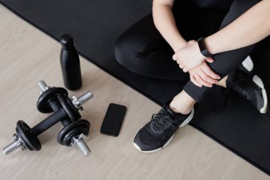spor ve spor geçmişi - siyah yoga minderinde oturan siyah ekipmanlı bir kadın - halterler, bir şişe su ve akıllı telefon