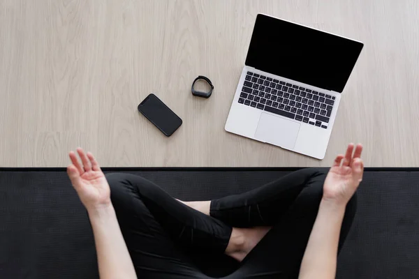 sport and technology concept - top view of young woman sitting in lotus pose on yoga mat, laptop and smartphone with blank screens and fitness tracker on the floor