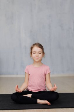 Evdeki yoga minderinde yoga egzersizleri yapan küçük tatlı bir kız. Gri duvar arkaplanının üzerinde boşluğu kopyalayan bir kız.