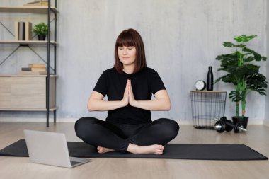 Genç ve güzel bir kadın evde oturmuş yoga egzersizleri yapıyor ve sanal dersleri laptopta öğreniyor.