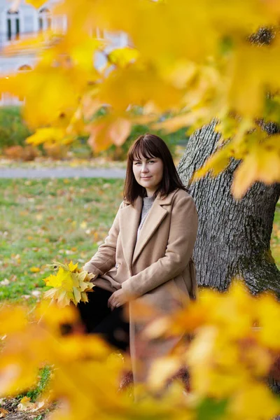 美丽而快乐的女人 身穿米黄色外套 坐在秋天的公园里 长着枫叶 — 图库照片