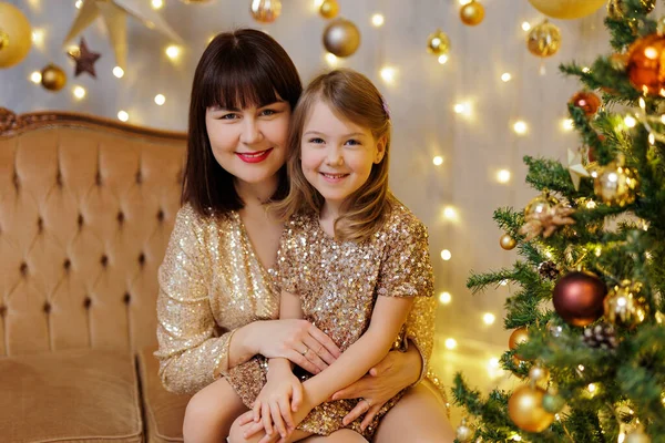 Mother and little daughter in golden dresses sitting on sofa near christmas tree