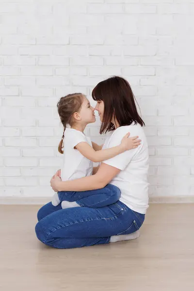 Happy mother and daughter sitting on the floor in jeans and white t-shirts near brick wall