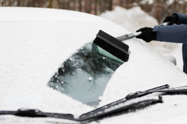 Adam arabasının ön camındaki karı büyük bir fırçayla temizliyor.