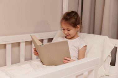 Küçük tatlı kız yatakta oturup kitap okuyor.