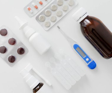 Dijital termometrenin üst görüntüsü, burun spreyi, haplar ve beyaz masadaki boğaz için sprey.