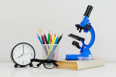 Mavi mikroskop, kitap, renkli kalemler ve beyaz masadaki saat.