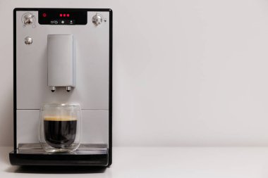 Masanın üzerinde cam kahve fincanı olan modern kahve makinesi ve duvardaki boşluğu kopyala.