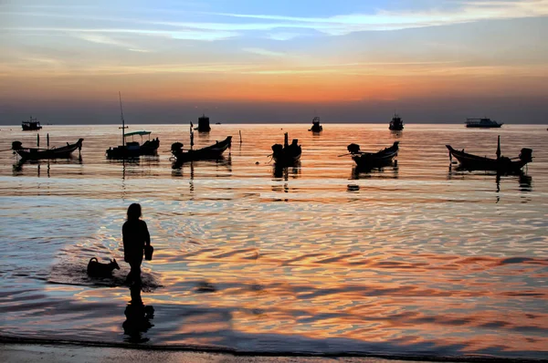 泰国高涛赛里海滩的长尾船与当地女孩和狗在日落时分的轮廓 图库图片