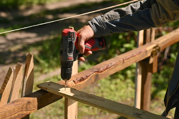 A man drills a hole in a wooden board with a drill. outdoors.
