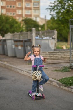 Küçük bir kız scooter kullanıyor. Bahçedeki çöp tenekelerinin yanında.