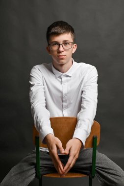 Gözlüklü bir çocuk gri arka planda bir sandalyede oturuyor.