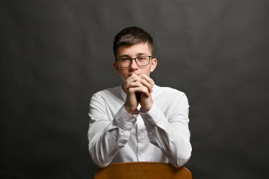 Parmaklarını önünde kenetle. Gri arka planda gözlüklü bir gencin portresi.