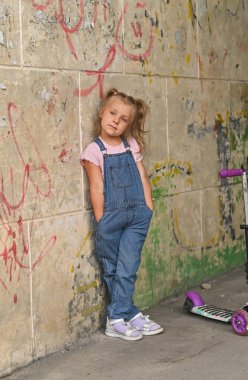 A little girl with her hands in her pants pockets poses against a shabby wall clipart