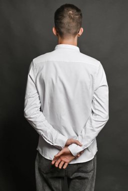 Dikey bir fotoğrafçının elleri arkadan bağlanmış, bir eli diğerinin bileğine dolanmış. Görüntü, gri arka planda bir gerilim, sabır ve içgözlem hissi veriyor..