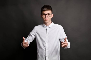 Beyaz gömlekli ve gözlüklü kendine güvenen genç adam, iki eliyle baş parmak kaldırıyor, basit gri arka planda. Fotoğraf onay, başarı ve olumlu tutumu simgeler, iş kavramları için mükemmeldir.