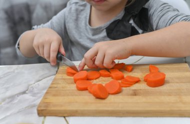 Turuncu havuç dilimleriyle kaplı ahşap kesme tahtası çocukların aşçılık gelişimini gösteriyor. Aile yemekleri ya da mutfak eşyaları reklamları için mükemmel bir fotoğraf. Çocuk mutfak bıçağıyla kesmeyi öğreniyor.