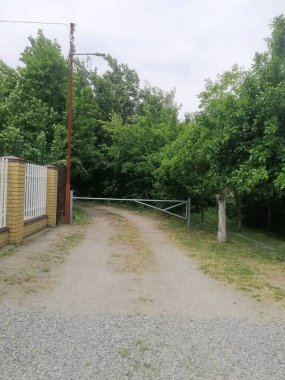 dikey fotograf. Kır yolu yakınındaki basit kapı ve çitlerle kırsal alanda sakin bir yaşam alanı yaratıyor. Doğal ortamdaki sarı tuğlalı çitin yanına açılan kapı mahremiyeti ifade ediyor.