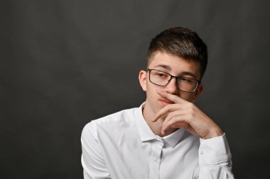 Gözlüklü düşünceli genç adam, gri stüdyo arka planına karşı ağzını eliyle kapatıyor. Düşünceli ifade derin bir konsantrasyon ve derin bir tefekkür gösteriyor. Düşünme konsepti. Boşluğu kopyala