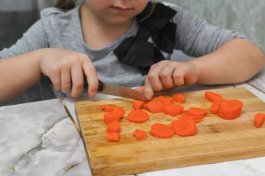 Küçük kız, ahşap tahtada pişirilmiş havuçları dikkatlice dilimliyor ve temel mutfak becerilerini öğreniyor. Çocuklar için bir ev mutfağında konsept pişirme dersi