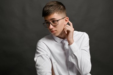 Gözlüklü çocuk önüne bakıyor, kulağının arkasını tırmalıyor, ne cevap vereceğini düşünüyor. Düşünceli ve düşünceli bir davranış. Tarafsız ifadeli, resmi tişörtlü genç bir çocuk. Düşünce anı.. 