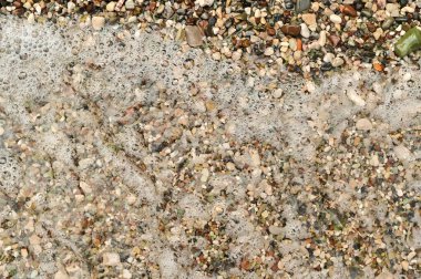 Çeşitli küçük taşlar, deniz kabukları ve ıslak kum tarafından yaratılmış soyut desen. Deniz kıyısında köpük köpüklü deniz çakıl taşlarının arka planı, doğal düzen. Dalga taşlı kıyı şeridiyle birleşiyor. 