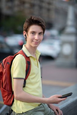Kulaklıklı, akıllı telefon kullanan, yaz parkında yaşayan yakışıklı bir çocuk. 15 yaşında bir genç konuşuyor ve cep telefonuyla mesajlar gönderiyor, gençlik yaşam tarzı.