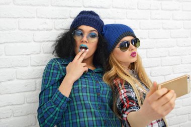 İki mutlu komik kız beyaz tuğlaların üzerinden akıllı telefonlarıyla selfie çekiyor.