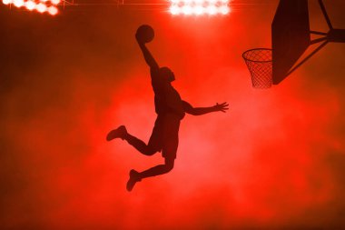 Genç profesyonel basketbolcunun gölgeli gölgesi koyu kırmızı duman arka planına smaç bastı