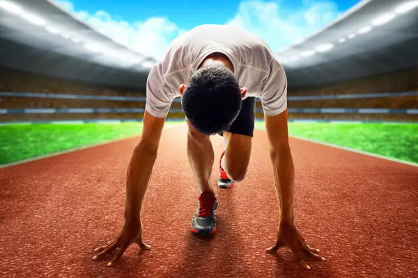 Asyalı genç adam yeşil çimenler ve kırmızı asfalt ile stadyum sahasında başlangıç pozisyonunda.