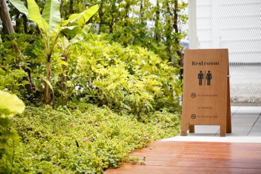 Yeşillik ve bitkilerle çevrili yemyeşil bahçe ortamındaki tuvalet tabelası sakin bir atmosfer yaratıyor. İşaretler sigara içilmediğini gösteriyor..