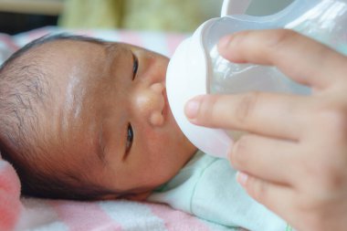 Yumuşak bir battaniyeye uzanmış yeni doğmuş bir bebek, bakıcı eliyle şişeden süt içiriliyor, şefkatli bir bakım ve beslenme sergiliyor.