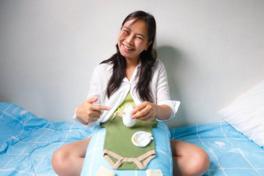 A smiling woman sitting on bed demonstrates baby care essentials, including green baby onesie, diapers, and baby bottle, creating warm and nurturing atmosphere clipart