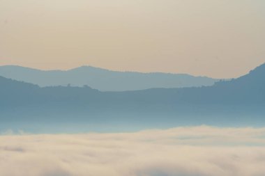 Yumuşak, katmanlı dağların sisle kaplandığı sakin bir manzara, sakin bir atmosfer yaratıyor. Mavi ve gri renkler huzur ve sükuneti çağrıştırıyor.