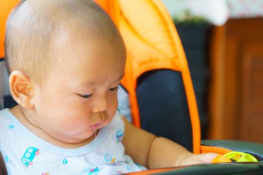 Odaklanmış bir bebek turuncu sandalyede oturuyor, merakla oyuncakları keşfediyor. Çocuğun kafası kel ve açık mavi gömlek giyiyor. Renkli desenleri var.