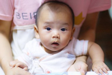 Geniş gözlü meraklı bir bebek kucağında oturup masumiyetini ve merakını gözler önüne seriyor. yumuşak ışıklandırma bebek özelliklerini ön plana çıkarıyor, sıcak ve yumuşak atmosfer yaratıyor