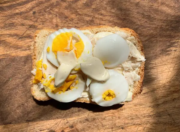 Bu yakın çekim, tahta kesme tahtasındaki açık yüzlü sandviçi gösteriyor. Bir dilim ekmek üzerine kremsi bir serpiştirme ve haşlanmış yumurta dilimleri ile birlikte ufalanmış yumurta sarısı görünür. Aydınlatma doğal görünüyor, biraz shad döküm