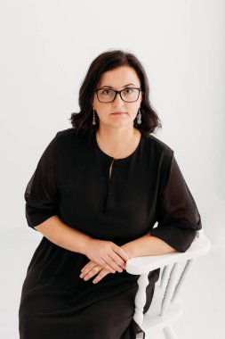 40 yaşında, kendine güvenen, zarif siyah bir elbise ve temiz beyaz bir arka planda gözlük takan bir kadının stüdyo portresi. Profesyonellik ve entelektüellik sergiliyor, imajı iş, şirket, moda ve yaşam tarzı için ideal kılıyor.