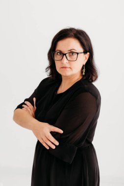 40 yaşında, kendine güvenen, zarif siyah bir elbise ve temiz beyaz bir arka planda gözlük takan bir kadının stüdyo portresi. Profesyonellik ve entelektüellik sergiliyor, imajı iş, şirket, moda ve yaşam tarzı için ideal kılıyor.