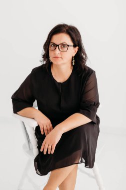 40 yaşında, kendine güvenen, zarif siyah bir elbise ve temiz beyaz bir arka planda gözlük takan bir kadının stüdyo portresi. Profesyonellik ve entelektüellik sergiliyor, imajı iş, şirket, moda ve yaşam tarzı için ideal kılıyor.