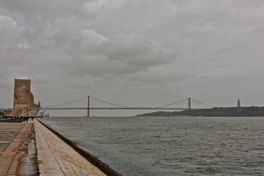 Lizbon, Portekiz 'deki 25 Nisan köprüsünün, soldaki Keşifler Anıtı' na bakan Tagus nehrinin üzerinde ve sağdaki Kral İsa 'ın Sığınağı' na uzaktan bakınız.