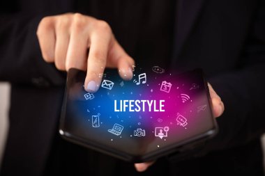 İş adamı, üzerinde LIFESTYLE yazıları ve sosyal medya kavramı olan katlanabilir bir akıllı telefon tutuyor.