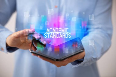 ACADEMIC STANDARDS yazıtlı, eğitici konseptli katlanabilir bir akıllı telefon tutan genç adam