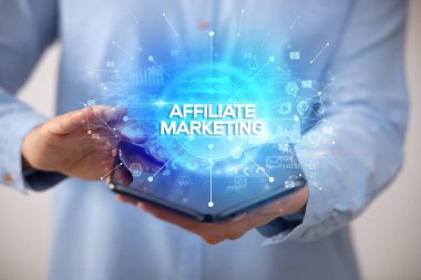 İş adamı elinde katlanabilir bir akıllı telefon tutuyor. Üzerinde AFFILIATE MarKETING yazıları ve yeni iş konsepti var.