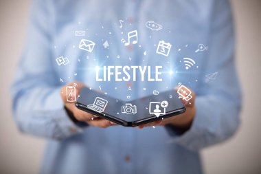 İş adamı, üzerinde LIFESTYLE yazıları ve sosyal medya kavramı olan katlanabilir bir akıllı telefon tutuyor.