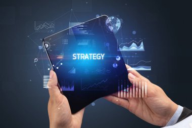 İş adamı elinde Strateji yazıtlı, başarılı bir iş konsepti olan katlanabilir bir akıllı telefon tutuyor.