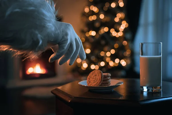 Weihnachtsmann Greift Gemütlichen Weihnachtszimmer Nach Dem Glas Milch Und Plätzchen lizenzfreie Stockfotos