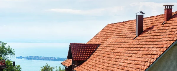 Chimneys on red tiled roof of residential house overlooking lake in summer. Red rooftop with metal and brick chimneys pipes on sky background. Banner with modern air vent system and roof tiles.
