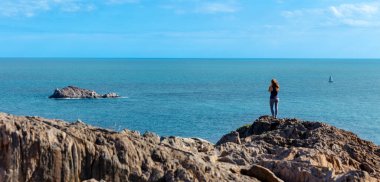 Kayanın üzerinde durup fotoğraf çeken ve güzel okyanus manzarasının tadını çıkaran bir kadın.