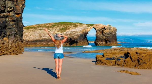 stock image Woman with arms raised enjoying atlantic ocean and rock formation- cathedral beach with arch and cave- Galicia,  Spain